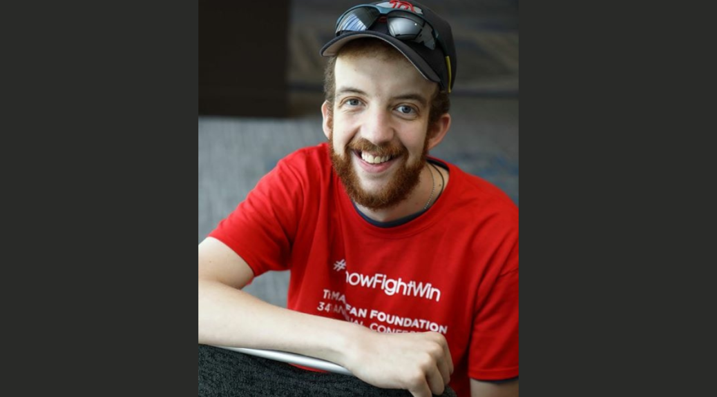 a young, caucasian man with brownish red hear and beard, wearing a red Marfan Foundation #KnowFightWin shirt, ball cap, and sunglasses, smiles at the camera.