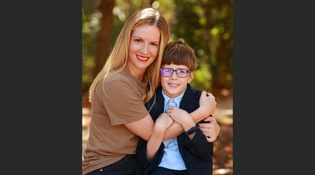 A blonde woman with straight hair hugs her 8 year old son, who has glasses and short brown hair.