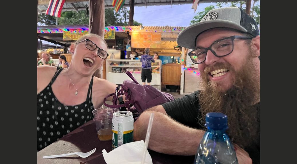 left- woman in a black tank top and glasses laughs uncontrollably. right- man with long brown beard, glasses, and hat laughs and smiles at camera