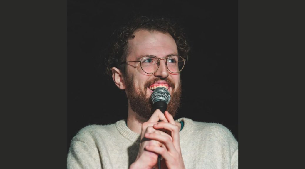tall white man with dark curly hair and glasses holds a microphone and smiles