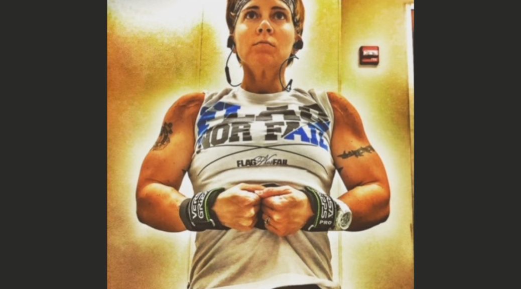muscular white woman in white tank top, wearing headphones, poses with her fists together and her muscles flexed