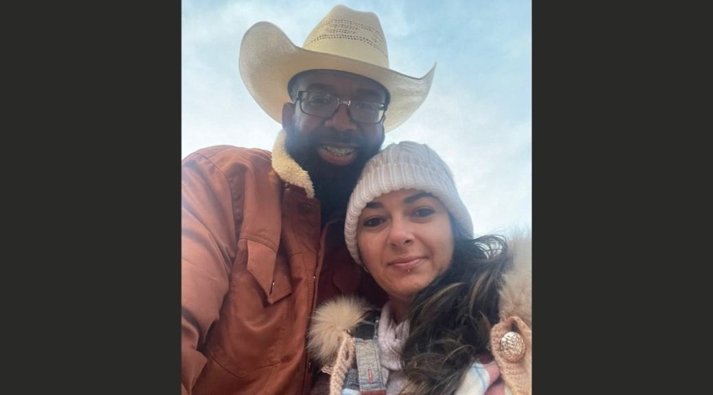 Black man with beard, glasses, and straw hat smiles with his arm around a woman with a white beanie and dark brown hair.