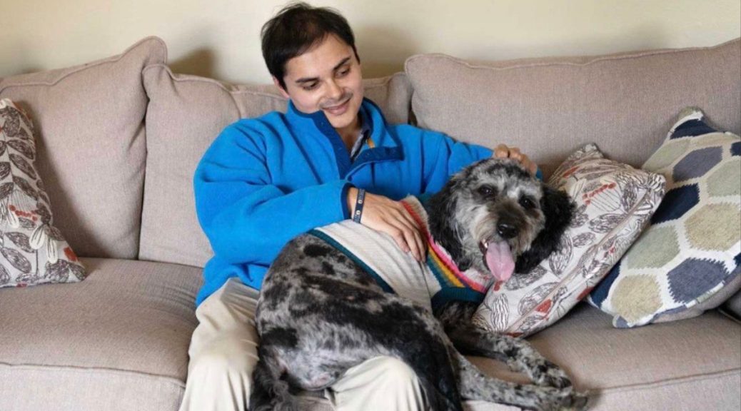Young man with dark hair, blue jacket, and khaki pants sits on a couch smiling at his dog, who is sitting on his lap. The dog is medium size, gray and black.